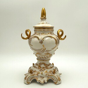 May include: A white and gold decorative urn with a lid. The urn features ornate gold scrollwork and handles. The base is also decorated with gold accents. The finial on the lid is gold. The urn is likely made of porcelain or ceramic.