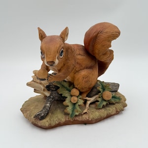 May include: A detailed figurine of a squirrel holding an acorn, perched on a branch with leaves and acorns. The squirrel has reddish-brown fur and a bushy tail. The base is a naturalistic design with green and brown tones.