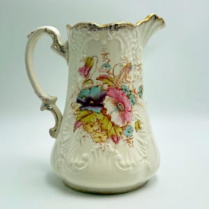 May include: Cream-colored ceramic pitcher with a floral design. The pitcher features a decorative handle and a gold-trimmed rim. The floral pattern includes pink, blue, and yellow flowers with green leaves. The pitcher has an embossed design.
