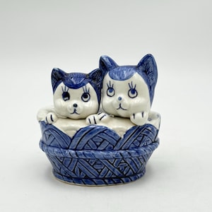 May include: A ceramic figurine featuring two stylized cats in a blue and white basket. The cats have blue ears and accents, with white faces and paws. The basket is intricately designed with a woven pattern.