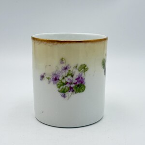 May include: White ceramic cup with a gold rim and a floral design. The cup features a cluster of purple violets with green leaves. The top of the cup has a gradient of beige to white.