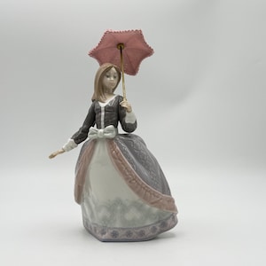May include: A porcelain figurine of a figure with long brown hair wearing a dark brown bodice and a voluminous skirt with white, grey, and pale pink panels. The figure holds a small, textured pink parasol with a gold-toned handle.