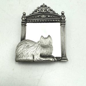 May include: A silver-toned cat-shaped mirror with an ornate frame. The cat is lying down, and the frame has a decorative top and column-like sides. The mirror is rectangular.