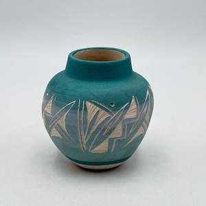 May include: A teal ceramic vase with a round body and a short neck. The vase features a carved design of stylized leaves and geometric shapes in a lighter color. The base is a lighter shade of teal.