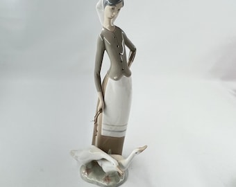 May include: A porcelain figurine of a woman in a long skirt and a headscarf, holding a small bundle. Two white geese are at her feet. The woman's outfit is gray and white. The base is a light green color.