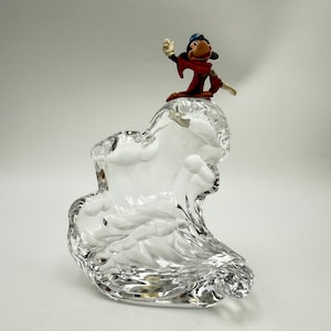 Op de afbeelding: Een heldere glazen sculptuur in de vorm van een golf, bekroond met een klein beeldje van een stripfiguur in een rode jurk en blauwe hoed. Het beeldje zwaait. De sculptuur staat tegen een witte achtergrond.