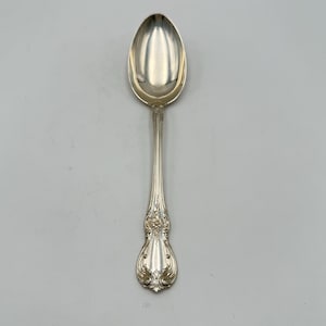 May include: A silver spoon with an oval bowl and a long, ornate handle. The handle features floral and scroll detailing. The spoon is set against a plain, light gray background, highlighting its polished surface and intricate design.