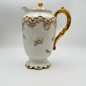 May include: A white porcelain coffee pot with a gold handle and accents. The pot features a floral design with pink flowers and gold trim. The lid is topped with a gold finial. The pot is approximately 10 inches tall.