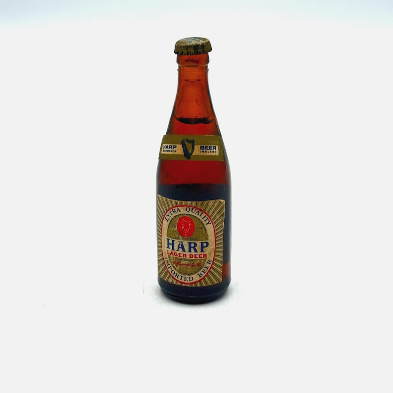 May include: A vintage Harp Lager Beer bottle with a brown glass body and a gold cap. The label features the Harp logo and the words "Extra Quality" and "Imported Beer". The bottle is likely from Ireland.