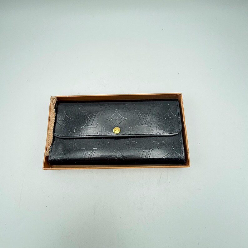 Puede incluir: Cartera de cuero negro con patr&oacute;n de monograma en relieve, asegurada por una solapa y un cierre de bot&oacute;n a presi&oacute;n dorado. La cartera se presenta dentro de una caja de cart&oacute;n, lo que sugiere un estado nuevo o sin usar.
