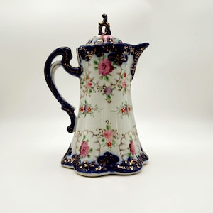 May include: A vintage porcelain coffee pot with a white base, adorned with pink roses and gold accents. The pot features a navy blue handle and trim, and a matching lid. The design is ornate and floral, with a scalloped base.