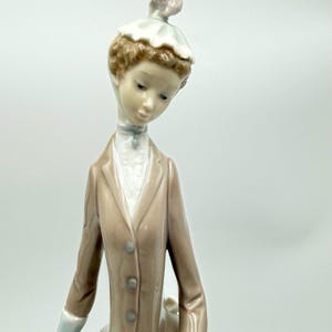 May include: A porcelain figurine of a woman in a long tan coat and a white hat with a pink accent. The figurine has a delicate face and is looking down. The coat has three buttons and the figurine is wearing a white shirt.