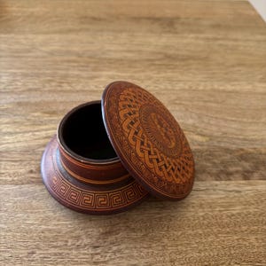 May include: A small, round, wooden box with a lid. The box is dark brown with intricate carved designs on the lid and base. The lid is slightly ajar, revealing the dark interior of the box. The design features geometric patterns and a central circular motif.