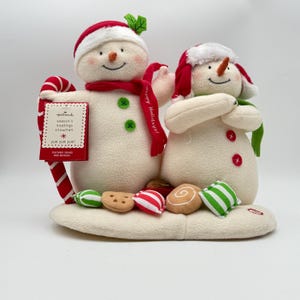 May include: A plush snowman duo with red and white hats and scarves. One holds a candy cane and a sign that reads "season's treatings snowmen." Festive candies and cookies are at the base. The text "Happy Holidays!" is on the scarf.