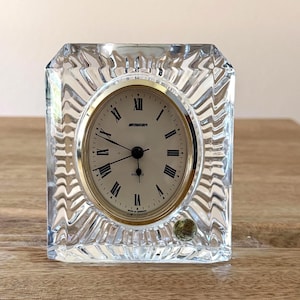 May include: A clear, square crystal quartz clock with a gold-tone face and black Roman numerals. The clock face reads "Stocker" and "Quartz". The clock has a decorative, ridged crystal frame.