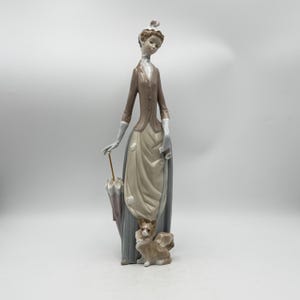 May include: A porcelain figurine of a woman in a long dress, holding an umbrella. The figure is in shades of brown, beige, and gray, with a small dog at her feet. The figurine has a classic, elegant design.