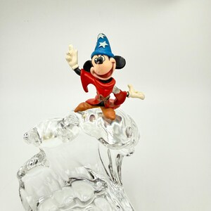 Op de afbeelding: Een beeldje van Mickey Mouse in een tovenaarsoutfit, met een blauwe hoed en rode mantel, bovenop een heldere, golfvormige glazen sculptuur. Het figuur staat in een dynamische pose, met &eacute;&eacute;n arm omhoog.