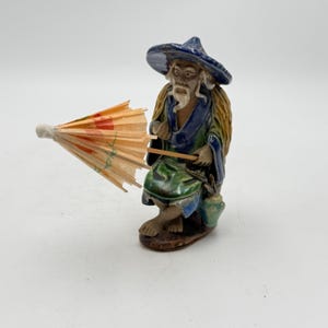 May include: A ceramic figurine of an elderly Asian man wearing a blue hat and robe, holding a small paper parasol. The figurine has a detailed face and is seated, with a small bucket beside him. The colors include blue, green, brown, and orange.