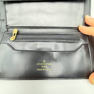 Puede incluir: Cartera de cuero negro con cremallera dorada y logotipo dorado. La cartera tiene el texto "LOUIS VUITTON PARIS made in France" impreso en dorado. La cartera est&aacute; abierta, revelando m&uacute;ltiples compartimentos.