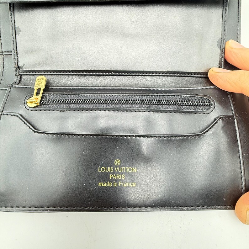Puede incluir: Cartera de cuero negro con cremallera dorada y logotipo dorado. La cartera tiene el texto "LOUIS VUITTON PARIS made in France" impreso en dorado. La cartera est&aacute; abierta, revelando m&uacute;ltiples compartimentos.