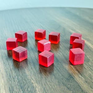 May include: A collection of ten translucent red dice, each cube-shaped with rounded edges. The dice are scattered on a dark wooden surface, with light reflecting off their surfaces. The image is well-lit, highlighting the color and form of the dice.