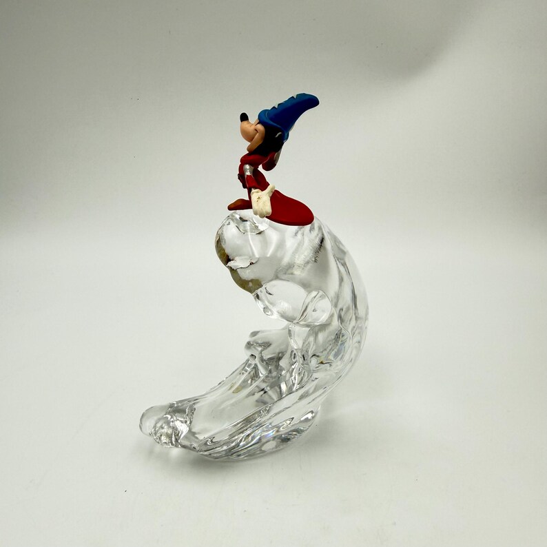 Op de afbeelding: Een beeldje van Mickey Mouse in een tovenaarsoutfit, bovenop een heldere, golfvormige glazen sculptuur. Mickey draagt een blauwe hoed, een rode mantel en witte handschoenen. De glazen basis heeft een glad, vloeiend ontwerp.