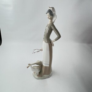 May include: A porcelain figurine of a woman in a long dress and a head covering, holding a small twig. The woman is standing next to two geese. The figurine is primarily white, gray, and beige.