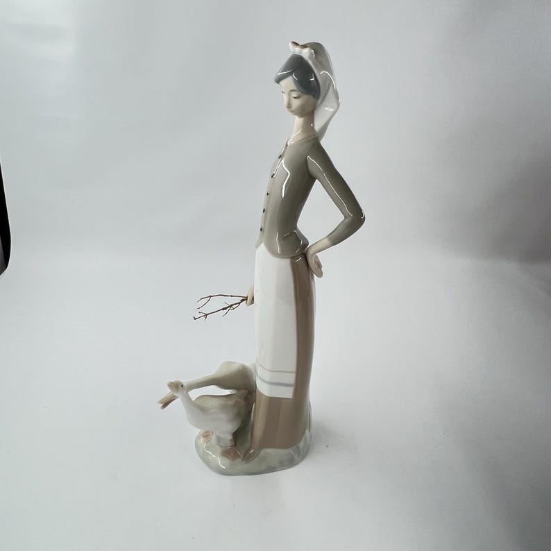 May include: A porcelain figurine of a woman in a long dress and a head covering, holding a small twig. The woman is standing next to two geese. The figurine is primarily white, gray, and beige.