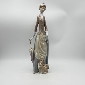 May include: A porcelain figurine of a woman in a long dress, holding an umbrella. The woman wears a brown jacket and a hat. A small dog figurine sits at her feet. The figurine is painted in muted tones of brown, beige, and gray.