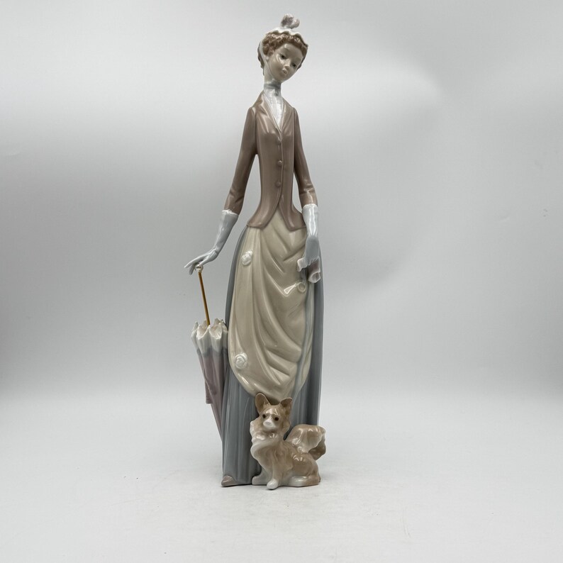 May include: A porcelain figurine of a woman in a long dress, holding an umbrella. The woman wears a brown jacket and a hat. A small dog figurine sits at her feet. The figurine is painted in muted tones of brown, beige, and gray.