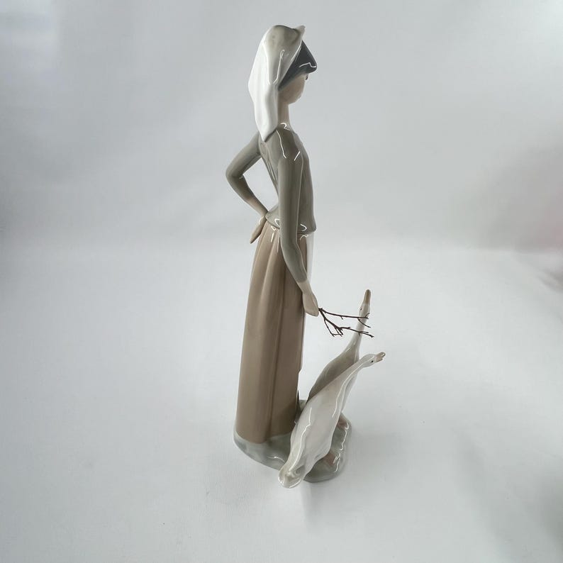 May include: A porcelain figurine of a woman in a long dress and head covering, holding a small branch. Two white geese stand at her feet. The figurine is primarily in neutral tones of beige, gray, and white.
