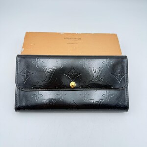 Puede incluir: Cartera de cuero negro con estampado floral y logotipo en relieve. La cartera tiene un cierre de bot&oacute;n dorado. El nombre de la marca "LOUIS VUITTON" es visible en la parte superior de la caja detr&aacute;s de la cartera.