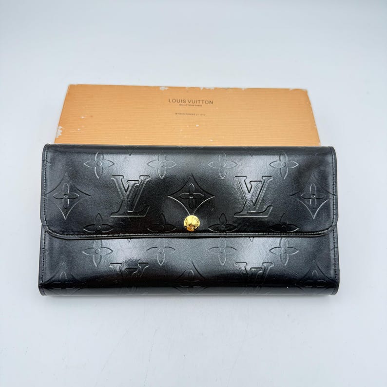 Puede incluir: Cartera de cuero negro con estampado floral y logotipo en relieve. La cartera tiene un cierre de bot&oacute;n dorado. El nombre de la marca "LOUIS VUITTON" es visible en la parte superior de la caja detr&aacute;s de la cartera.