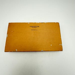 Puede incluir: Una caja rectangular de color marr&oacute;n claro con una superficie texturizada. La caja tiene las palabras "LOUIS VUITTON MALLETIER PARIS" y "MAISON FOND&Eacute;E EN 1854" impresas en la parte superior. Los bordes de la caja muestran signos de desgaste.