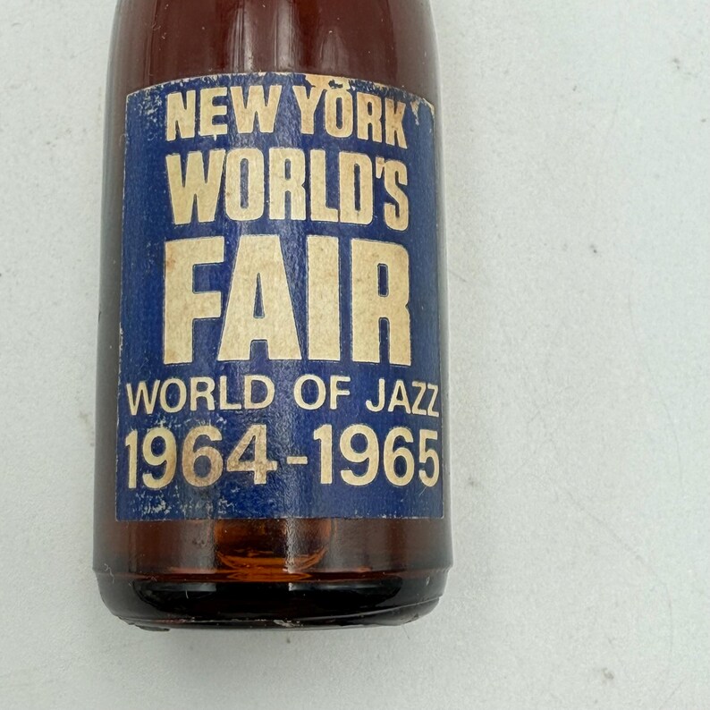 May include: A brown glass bottle with a blue and white label. The label reads "NEW YORK WORLD'S FAIR" with "WORLD OF JAZZ 1964-1965" below. The bottle is partially filled with a dark liquid.