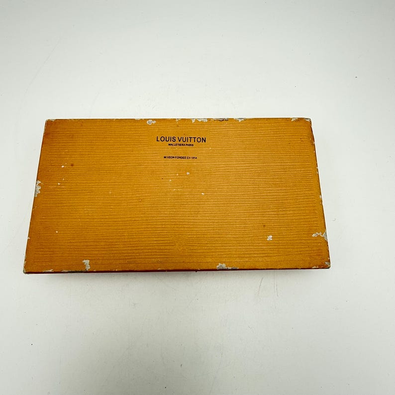 Puede incluir: Una caja rectangular de color marr&oacute;n claro con el texto "LOUIS VUITTON MALLETIER PARIS" impreso en la parte superior. La caja tiene una superficie texturizada con l&iacute;neas horizontales y algo de desgaste en los bordes. Tambi&eacute;n es visible el texto "MAISON FOND&Eacute;E EN 1854".