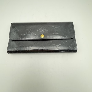 Puede incluir: Cartera de cuero negro con estampado de monograma en relieve. La cartera tiene un cierre de solapa con un bot&oacute;n dorado. El dise&ntilde;o incluye el logotipo de la marca y motivos florales, ofreciendo un estilo cl&aacute;sico y reconocible.