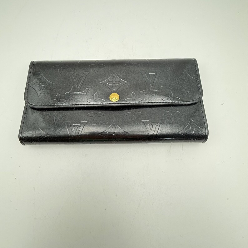 Puede incluir: Cartera de cuero negro con estampado de monograma en relieve. La cartera tiene un cierre de solapa con un bot&oacute;n dorado. El dise&ntilde;o incluye el logotipo de la marca y motivos florales, ofreciendo un estilo cl&aacute;sico y reconocible.