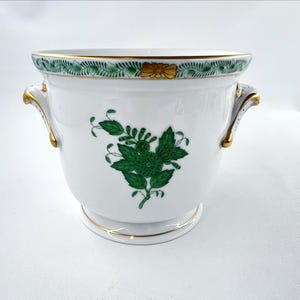 May include: White ceramic planter with green floral design and gold accents. The planter features two gold-trimmed handles and a decorative green and gold border. A detailed green floral pattern adorns the front of the planter.