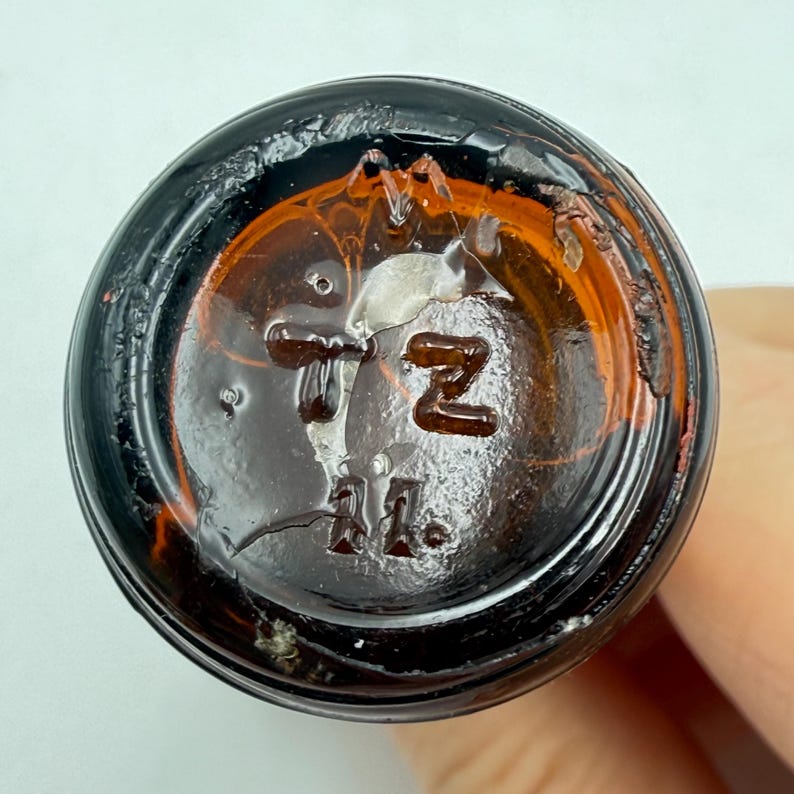 May include: Close-up of the bottom of a brown glass bottle. The base has embossed text including the letters "TZ" and the number "11". The glass has a dark amber color and shows signs of age.