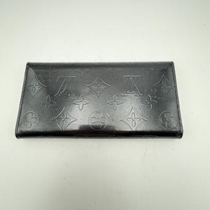 Puede incluir: Una cartera de cuero negro con patrones florales y logotipos en relieve. La cartera rectangular tiene un acabado brillante. El dise&ntilde;o incluye los motivos caracter&iacute;sticos de la marca, creando un accesorio de lujo.