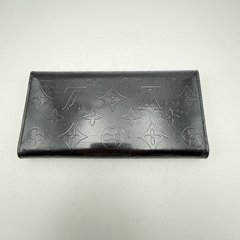 Puede incluir: Una cartera de cuero negro con patrones florales y logotipos en relieve. La cartera rectangular tiene un acabado brillante. El dise&ntilde;o incluye los motivos caracter&iacute;sticos de la marca, creando un accesorio de lujo.