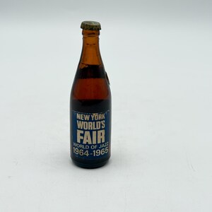 May include: A vintage brown glass bottle with a metal cap. The bottle has a blue and white label that reads "New York World's Fair" and "World of Jazz 1964-1965". The bottle is filled with a dark liquid.