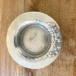 May include: A silver-colored, round coaster with a detailed grape and leaf design on the rim. The coaster has a slightly recessed center and a smooth, reflective surface. The coaster is likely made of metal.