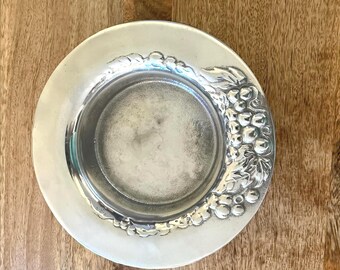 May include: A silver-colored, round coaster with a detailed grape and leaf design on the rim. The coaster has a slightly recessed center and a smooth, reflective surface. The coaster is likely made of metal.