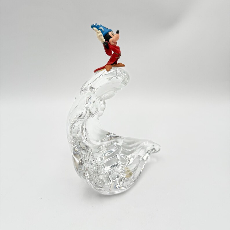 Op de afbeelding: Een heldere, gebeeldhouwde glazen figuur met een golfachtig ontwerp. Een kleine, kleurrijke Mickey Mouse-figuur in een tovenaarsoutfit staat erop. De achtergrond is wit.