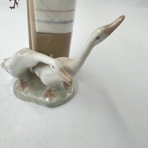 May include: A porcelain figurine of two geese, one with its head bent down, the other with its head and neck extended upwards. The geese are white with tan beaks and sit on a light green base with pink feet. A tall, brown and white striped vase is in the background.