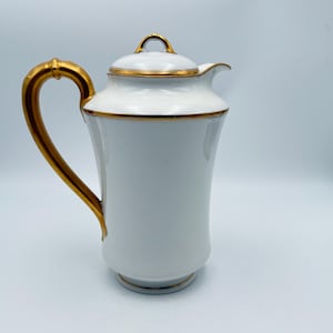 May include: White porcelain coffee pot with a gold handle and trim. The pot has a lid with a small loop handle and a spout for pouring. The gold accents provide a striking contrast against the white porcelain.