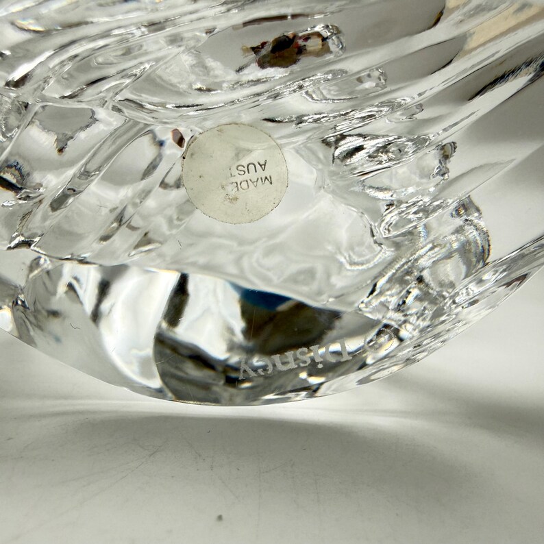 Op de afbeelding: Close-up van een helder, getextureerd glazen object met het woord "Disney" gegraveerd op de bodem. Een ronde sticker met de tekst "MADE AUST" is zichtbaar. Het object heeft een reflecterend, kristallijn uiterlijk.