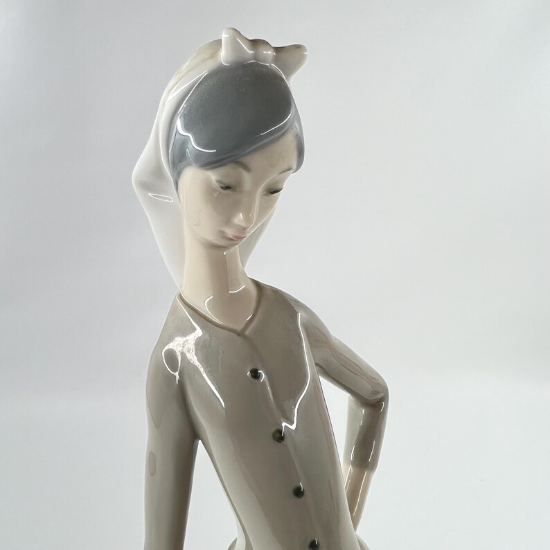 May include: A porcelain figurine of a woman in a long-sleeved, button-down dress. The figurine has a white head covering and gray hair. The woman is looking down with a slight smile. The figurine is a light gray color.